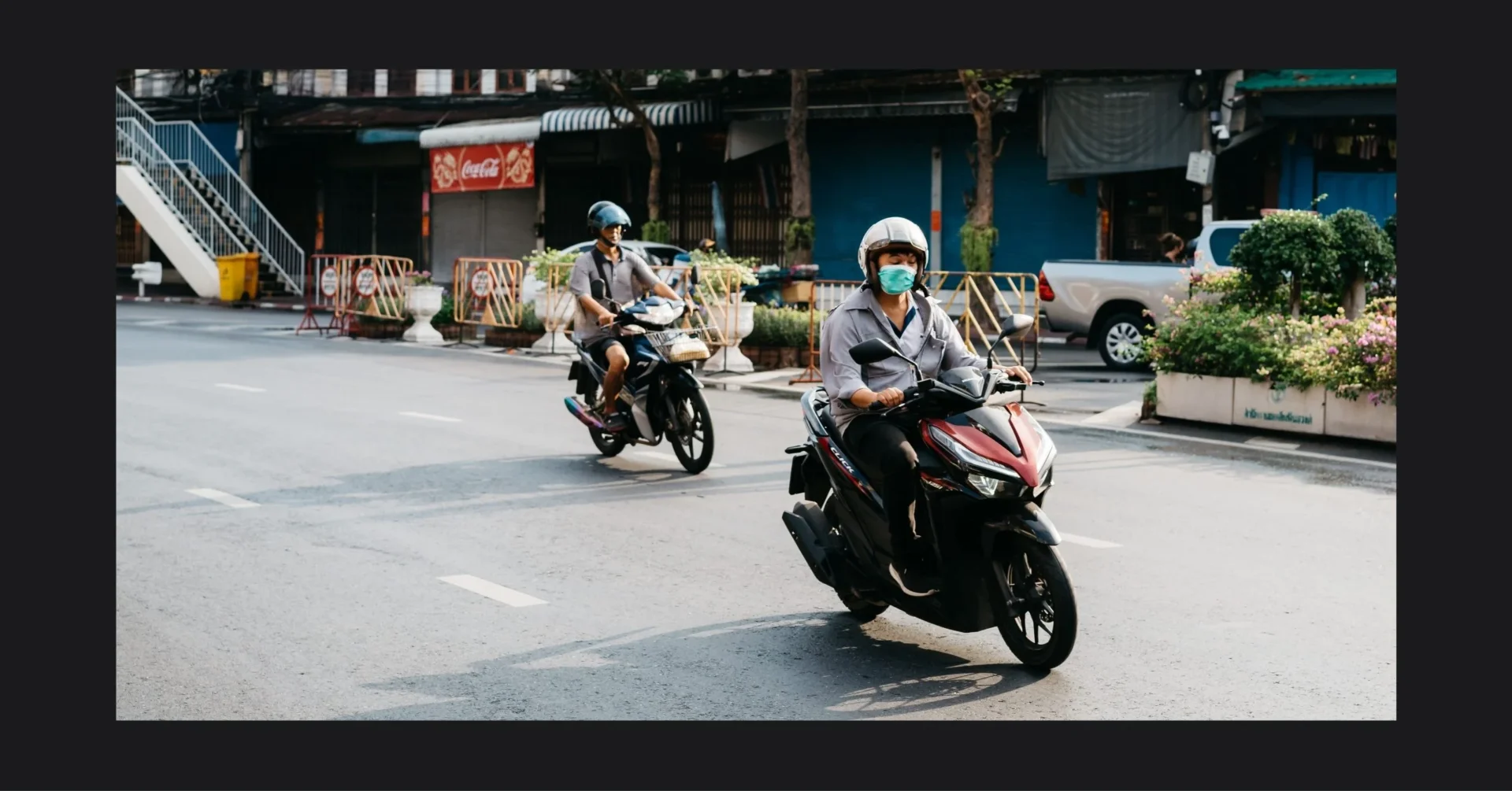 Two people riding motor scooters on a city street; both wear helmets and masks, with storefronts and construction barriers in the background.