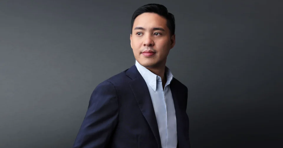 Portrait of a man in a navy suit and light blue shirt, looking to the right against a gray studio backdrop.